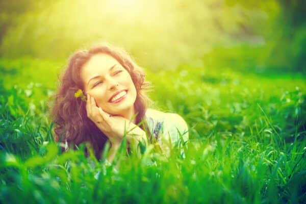 Peaceful looking woman lying in tall grass while holding a dandelion to her face and smiling.