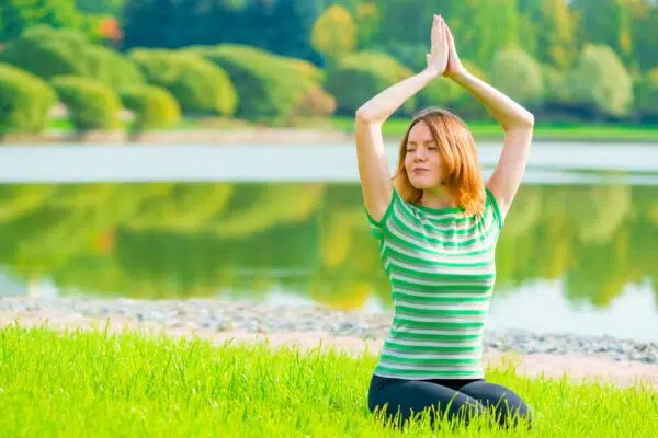 Young woman is sitting in the grass doing a yoga pose with her hands together over her head.