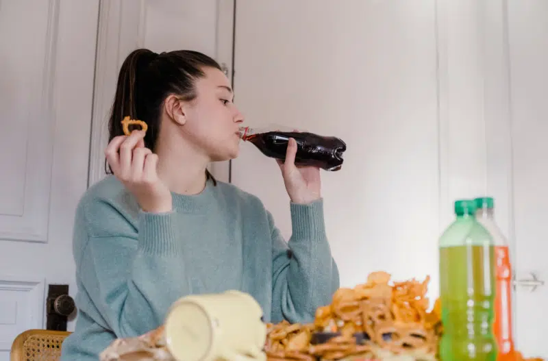 Woman is drinking from a soda bottle with a pretzel in her other hand.