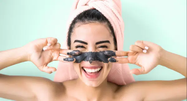 Smiling woman with a light pink bath towel wrapped around her head while rubbing a charcoal facemask across her nose and cheeks.