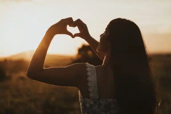 Woman forming a heart with her hands with the rising or setting sun in the background.